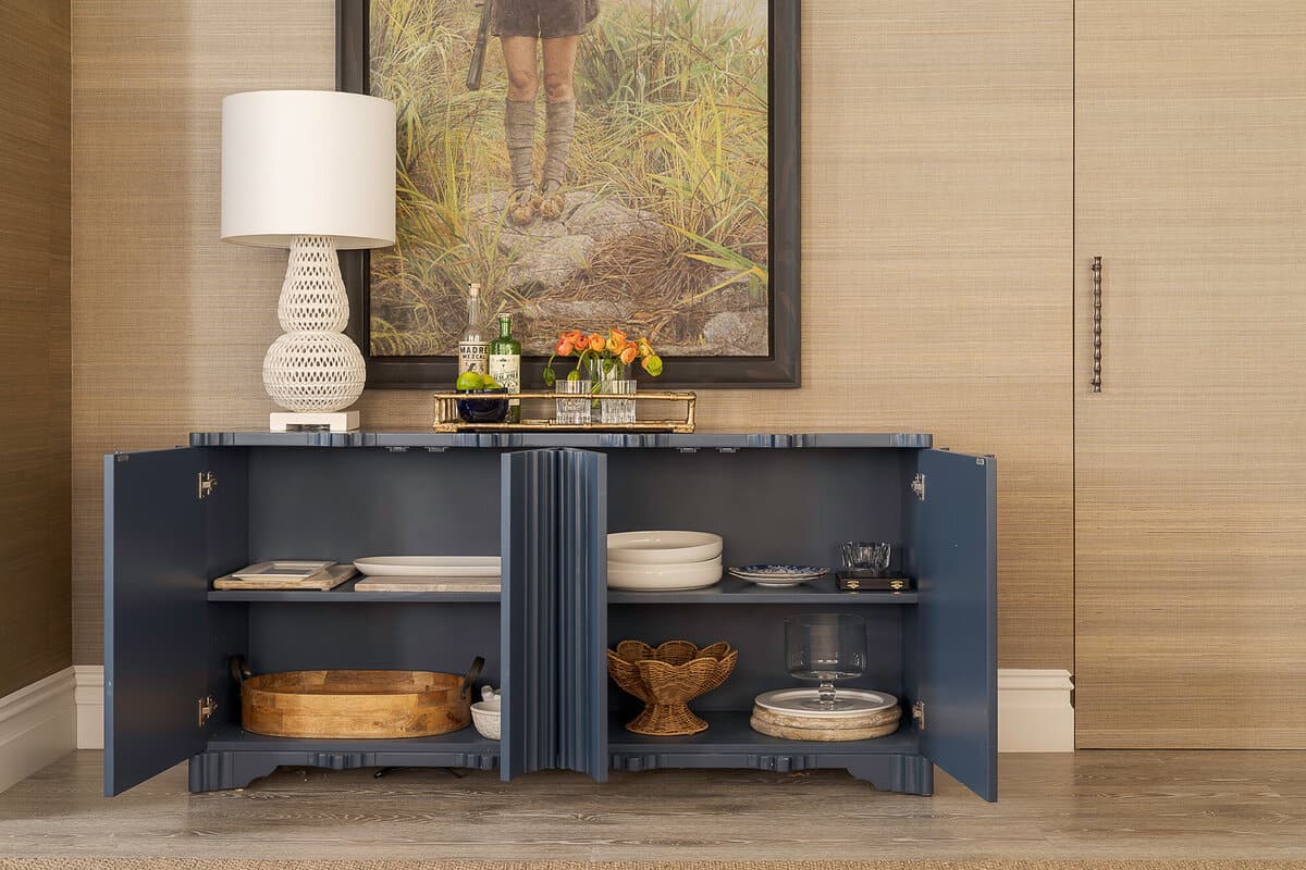 A navy fluted sideboard standing open against a grasscloth wall, holding stacked white plates, a wooden bowl and a woven fruit stand, with a white ceramic lamp and framed painting above.
