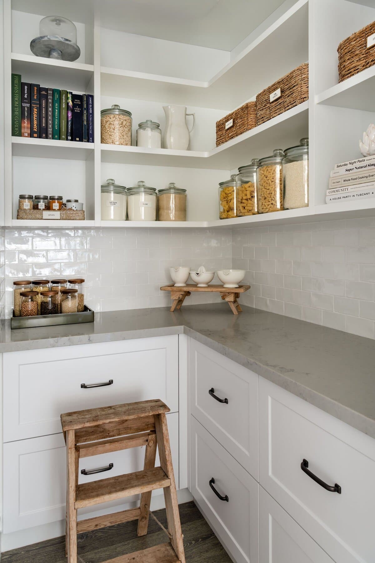 A pantry corner with glass jars of dry goods on the upper shelves, decanted spice jars, white crockery and a small timber stool below.