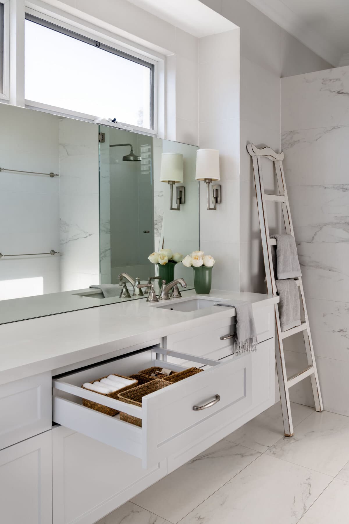 A bathroom vanity with a wide mirror, wall lights on either side, white flowers, an open drawer and a timber ladder holding a towel.