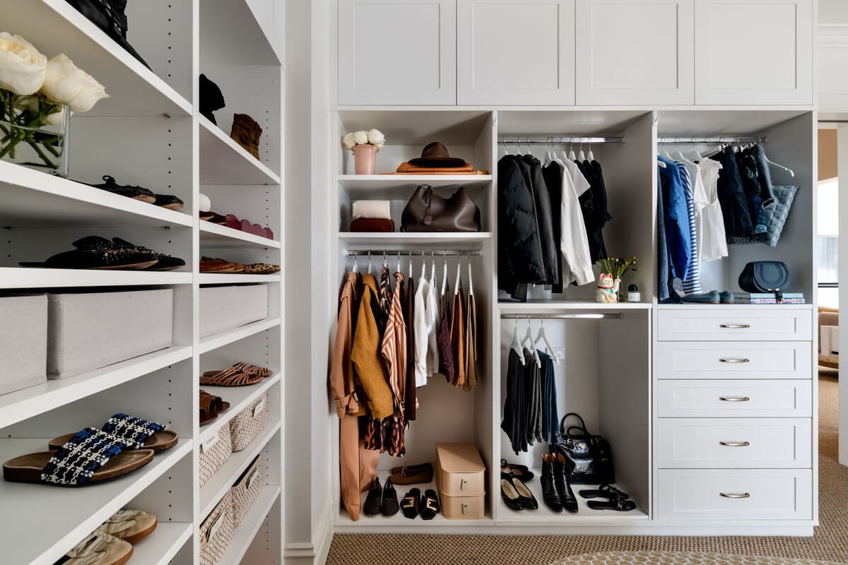 A walk in robe with open shelving, hanging clothes in rust, black and cream, and shoes lined up below.