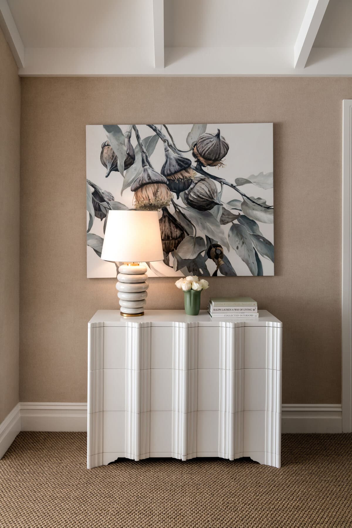 A framed botanical painting hanging above a fluted white console with a ribbed table lamp, against a grasscloth wall.