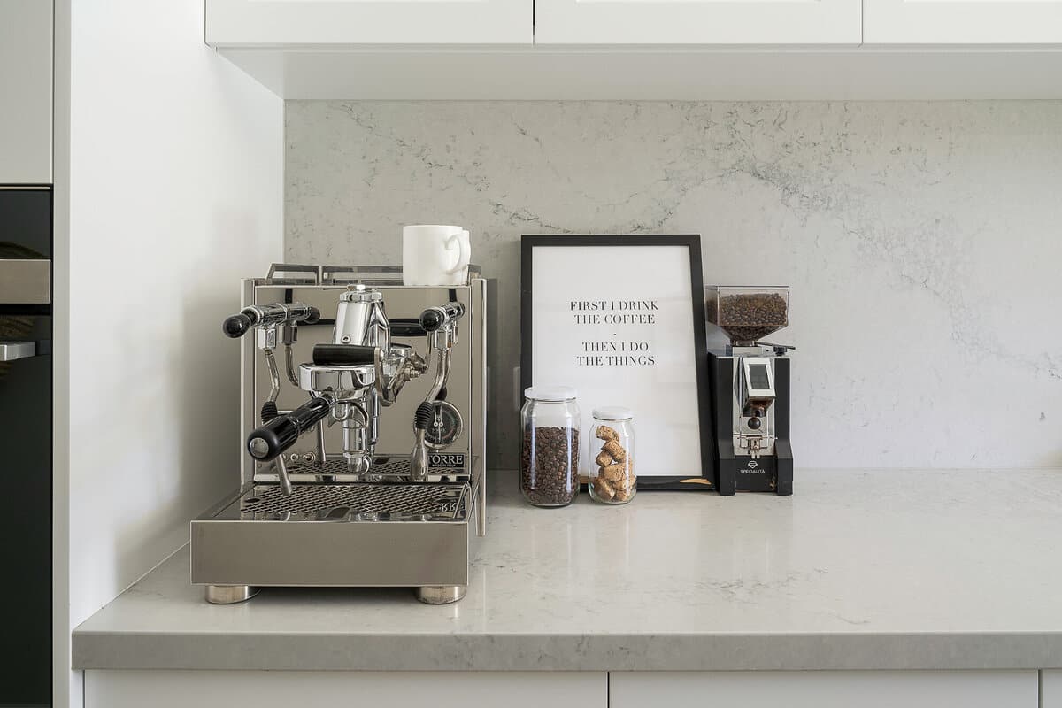 A stainless espresso machine on a marble benchtop beside a framed print and a glass jar of coffee beans.