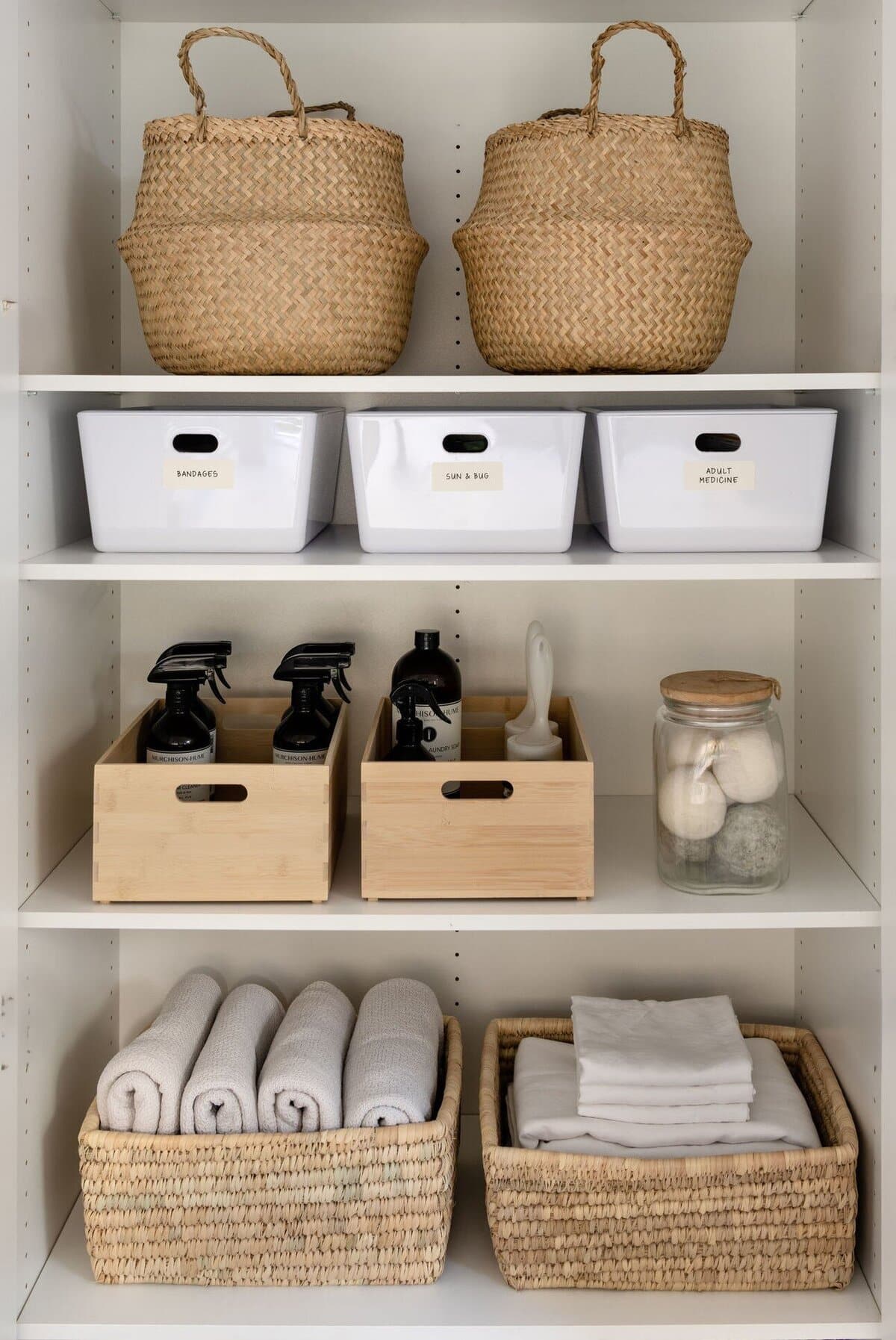 White laundry shelving with labelled tubs and decanted bottles on the upper shelf and rolled white towels in a timber crate below.